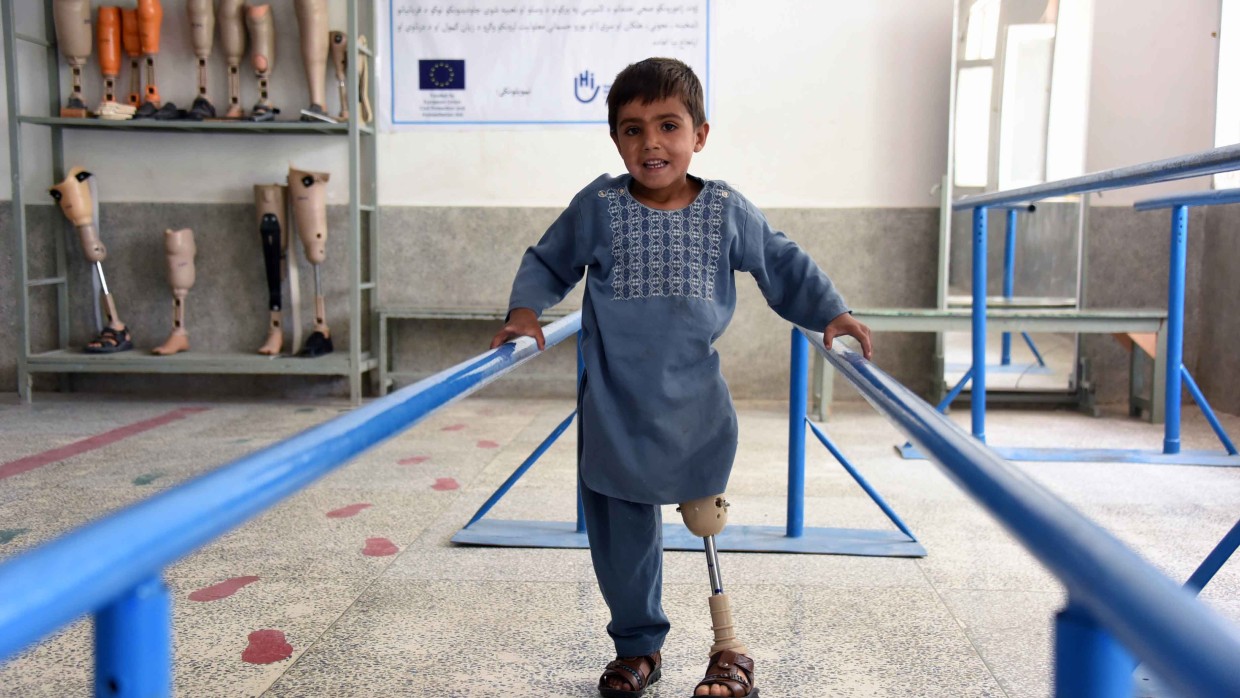 Ein Junge mit einer Beinprothese balanciert zwischen zwei blauen Handläufen in einem Rehabilitationszentrum. Im Hintergrund stehen Prothesen auf Regalen.