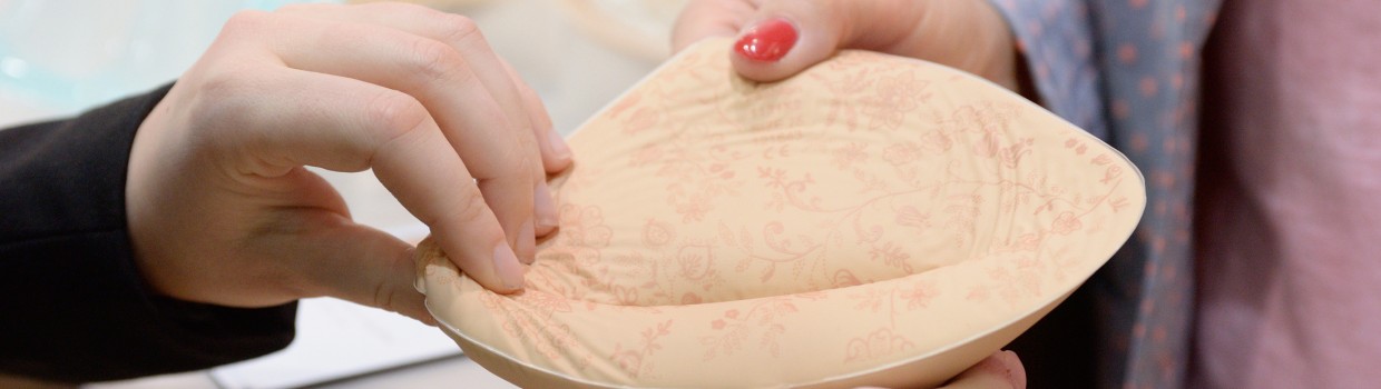 Zwei Personen halten gemeinsam eine Brustprothese aus weichem, hautfarbenem Material mit floralem Muster in den Händen. Zwei Personen halten gemeinsam eine Brustprothese aus weichem, hautfarbenem Material mit floralem Muster in den Händen.