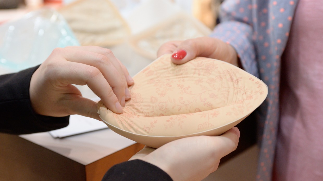 Zwei Personen halten gemeinsam eine Brustprothese aus weichem, hautfarbenem Material mit floralem Muster in den Händen.