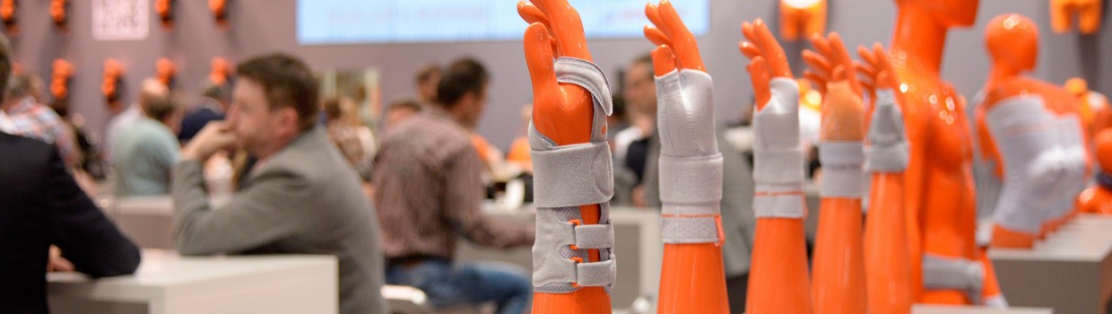 Mehrere orangefarbene Armprothesen mit grauen Orthesen sind in einer Reihe auf einem Tisch ausgestellt; im Hintergrund sitzen Menschen an Tischen, an der Wand hängen weitere orangefarbene Torso-Dummies und ein großer Bildschirm zeigt eine Präsentation. Mehrere orangefarbene Armprothesen mit grauen Orthesen sind in einer Reihe auf einem Tisch ausgestellt; im Hintergrund sitzen Menschen an Tischen, an der Wand hängen weitere orangefarbene Torso-Dummies und ein großer Bildschirm zeigt eine Präsentation.