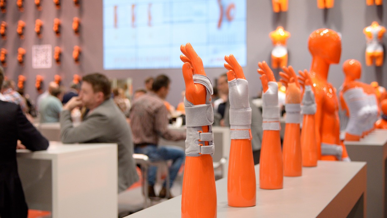Mehrere orangefarbene Armprothesen mit grauen Orthesen sind in einer Reihe auf einem Tisch ausgestellt; im Hintergrund sitzen Menschen an Tischen, an der Wand hängen weitere orangefarbene Torso-Dummies und ein großer Bildschirm zeigt eine Präsentation.
