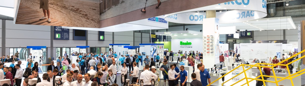 Messestand auf der OTWorld mit zahlreichen Besucherinnen und Besuchern, mehreren Exponaten wie Beinprothesen auf Tischen und großen Deckenbannern mit Fotos von Menschen mit Prothesen; im Hintergrund sind Stände von Unternehmen wie „DARCO“, „Basko“ und „Össur“ zu sehen.