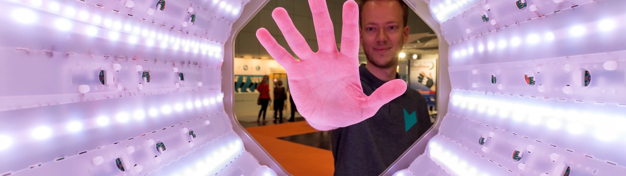 A hand holds five differently designed insoles fanned out at OTWorld; in the background, more insoles are laid out on tables and people are seen in conversation.