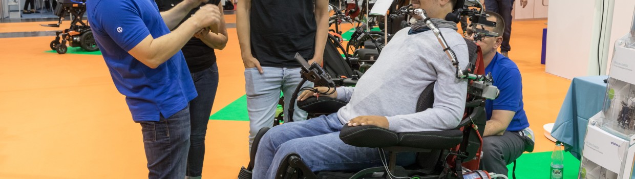 A woman demonstrates an ankle brace on a white model leg with a yellow sports shoe at the OTWorld booth; on the table are additional model arms and legs fitted with gray braces by the brand PUSH, with the text “ANKLE BRACE” visible in the background. A woman demonstrates an ankle brace on a white model leg with a yellow sports shoe at the OTWorld booth; on the table are additional model arms and legs fitted with gray braces by the brand PUSH, with the text “ANKLE BRACE” visible in the background.
