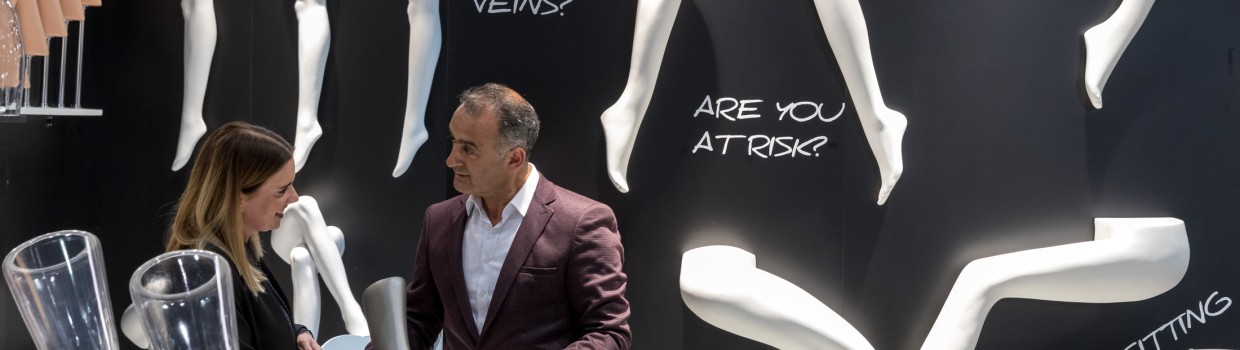 Zwei Personen stehen auf der OTWorld vor einer schwarzen Ausstellungswand mit weißen Modellbeinen und großem Text wie „VARICOSE VEINS?“ und „PROLONGED SITTING OR STANDING?“; sie betrachten gemeinsam ein Ausstellungsstück in Beinform. Zwei Personen stehen auf der OTWorld vor einer schwarzen Ausstellungswand mit weißen Modellbeinen und großem Text wie „VARICOSE VEINS?“ und „PROLONGED SITTING OR STANDING?“; sie betrachten gemeinsam ein Ausstellungsstück in Beinform.