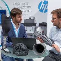 Zwei Männer sprechen am HP-Messestand auf der OTWorld über 3D-gedruckte Bauteile für Prothesen, darunter ein Beinprothesenschaft und gitterartige Strukturen; auf dem Tisch liegen weitere schwarze Demonstrationsobjekte, im Hintergrund sind Geräte und ein Bildschirm sichtbar.