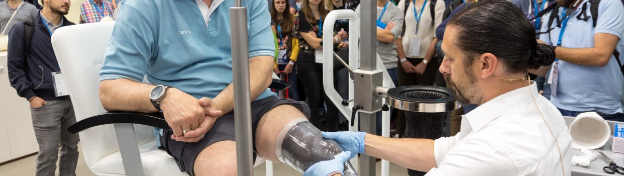 Ein Mann sitzt auf der OTWorld auf einem Stuhl, während ihm ein Orthopädietechniker einen durchsichtigen Guss für eine Beinprothese anpasst; zahlreiche Messebesucher beobachten die Demonstration im Hintergrund. Ein Mann sitzt auf der OTWorld auf einem Stuhl, während ihm ein Orthopädietechniker einen durchsichtigen Guss für eine Beinprothese anpasst; zahlreiche Messebesucher beobachten die Demonstration im Hintergrund.