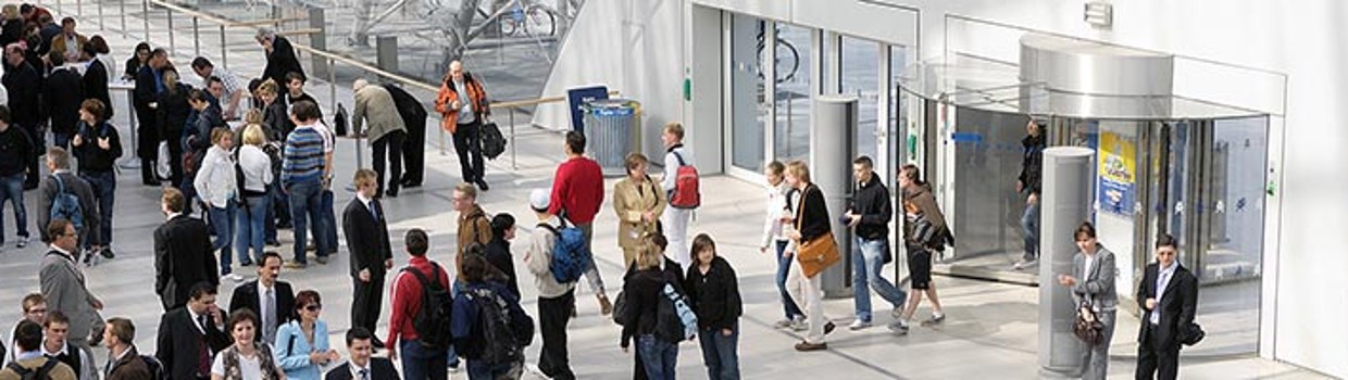 Menschen betreten und verlassen durch mehrere Drehtüren die Glashalle der Leipziger Messe; im lichtdurchfluteten Eingangsbereich herrscht reger Besucherverkehr.