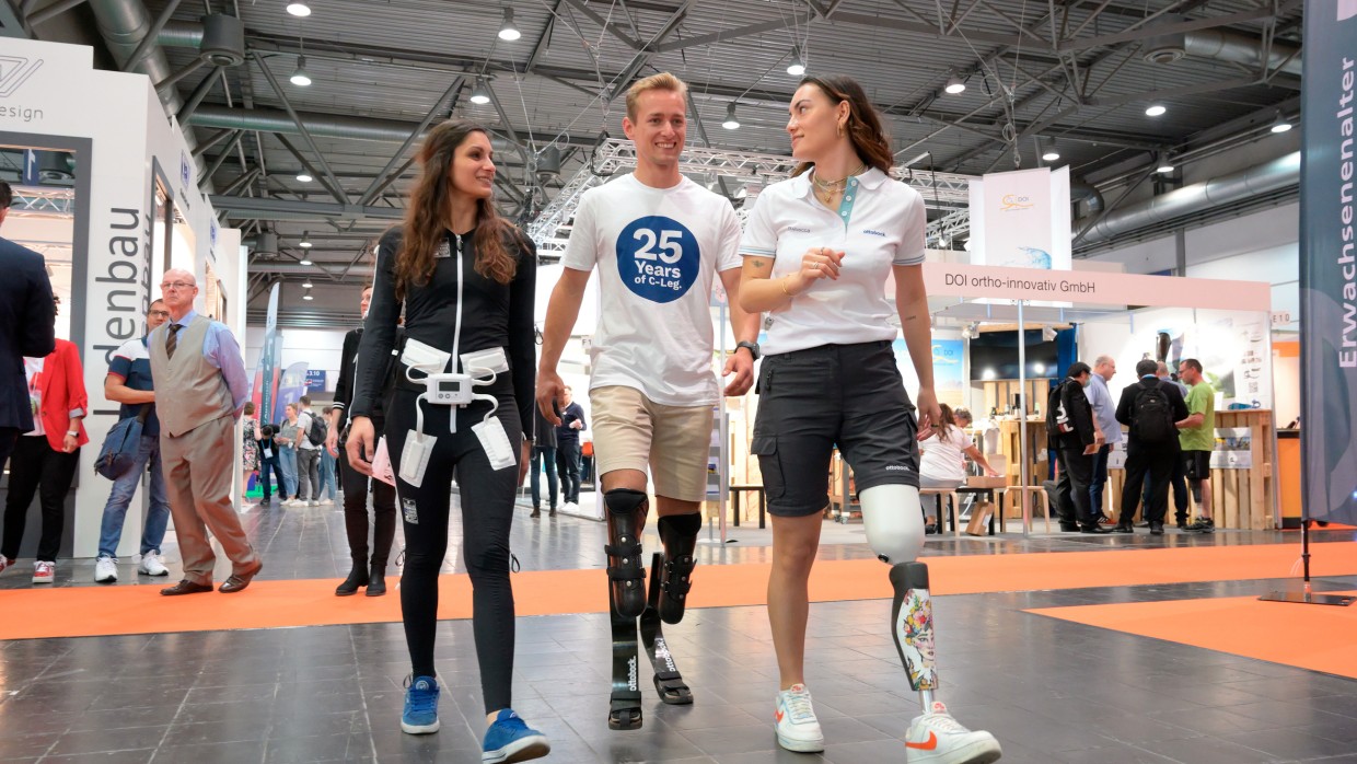 Drei junge Menschen mit verschiedenen orthopädischen Hilfsmitteln gehen lachend durch eine Halle der OTWorld. Drei junge Menschen mit verschiedenen orthopädischen Hilfsmitteln gehen lachend durch eine Halle der OTWorld.