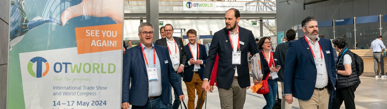 Gruppe von Messebesuchern betritt die OTWorld über die Glashalle. Links ist einPlakat mit Logo der OTWorld und Datum zu sehen. 