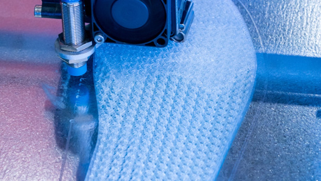 Ein 3D-Drucker druckt eine orthopädische Einlage aus hellem Material auf einer Metallplattform; im Bild sichtbar sind der Druckkopf mit Kabeln sowie das sich formende Objekt unter blauem Licht.