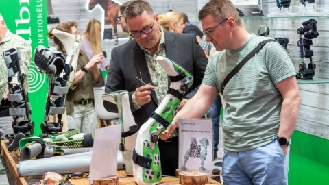 Zwei Männer begutachten auf der OTWorld kindgerechte Orthesen mit Fußballmotiven an einem Ausstellungsstand mit grünem Hintergrund.