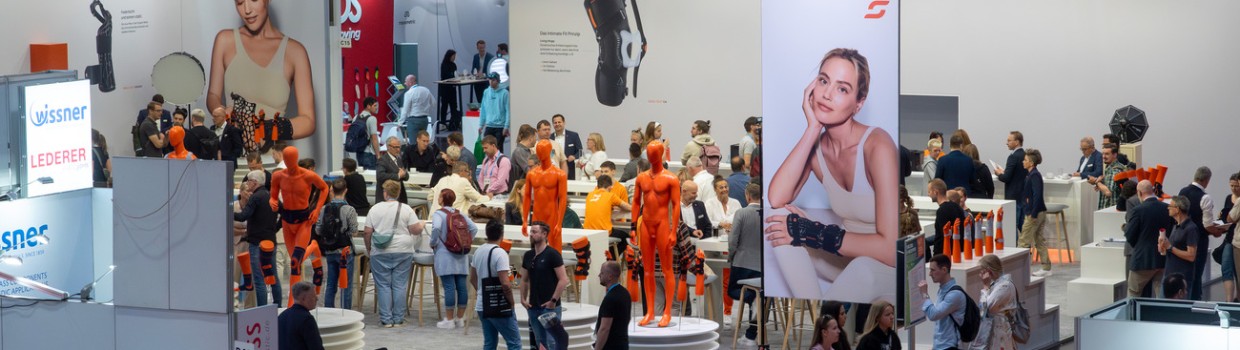 Großer Messestand von SPORLASTIC auf der OTWorld mit zahlreichen Besuchern, auffälligen orangen Schaufensterpuppen und großformatigen Produktplakaten. Großer Messestand von SPORLASTIC auf der OTWorld mit zahlreichen Besuchern, auffälligen orangen Schaufensterpuppen und großformatigen Produktplakaten.