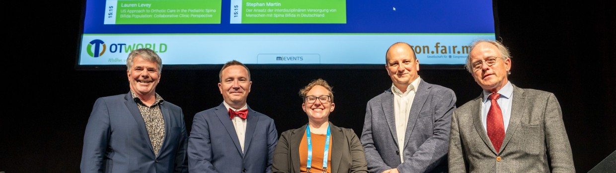 Fünf Personen stehen nebeneinander auf einer Bühne und blicken in die Kamera. Im Hintergrund ist eine große Leinwand mit Präsentationsfolien zur orthopädischen Versorgung zu sehen. Die Personen tragen Konferenz-Badges.