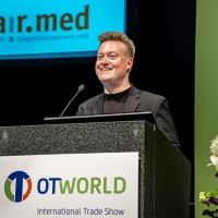 Ein Redner steht an einem Podium mit dem OTWorld-Logo und spricht auf der internationalen Fachmesse. Im Hintergrund ist das Logo der Firma "confairmed Gesellschaft für Congressmanagement mbH" zu sehen.