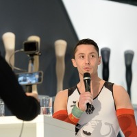 A man in a sports jersey with red compression sleeves speaks into a microphone; in the foreground, two people are filming with smartphones A man in a sports jersey with red compression sleeves speaks into a microphone; in the foreground, two people are filming with smartphones