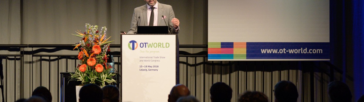 A man in a suit speaks on stage behind a podium with the "OTWorld" logo in front of an audience; to the left is a flower arrangement, and in the background are a screen with the "Hocoma" logo and a banner reading "Leipzig, Germany" and "www.ot-world.com". A man in a suit speaks on stage behind a podium with the "OTWorld" logo in front of an audience; to the left is a flower arrangement, and in the background are a screen with the "Hocoma" logo and a banner reading "Leipzig, Germany" and "www.ot-world.com".