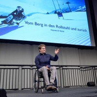 A man in a wheelchair is giving a talk on stage, with a large screen behind him showing an image of a monoski skier and the text “Vom Berg in den Rollstuhl und zurück – Keynote OT World”. A man in a wheelchair is giving a talk on stage, with a large screen behind him showing an image of a monoski skier and the text “Vom Berg in den Rollstuhl und zurück – Keynote OT World”.