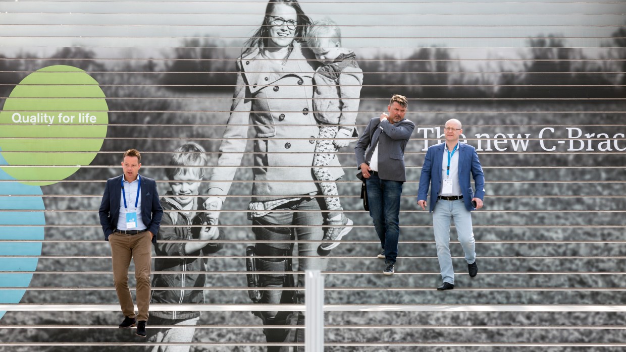 Three men walk down a large staircase with a photorealistic advertisement in the background showing a woman with two children in a landscape; the staircase displays the logos “ottobock.” and the slogans “Quality for life” and “The new C-Brace.”