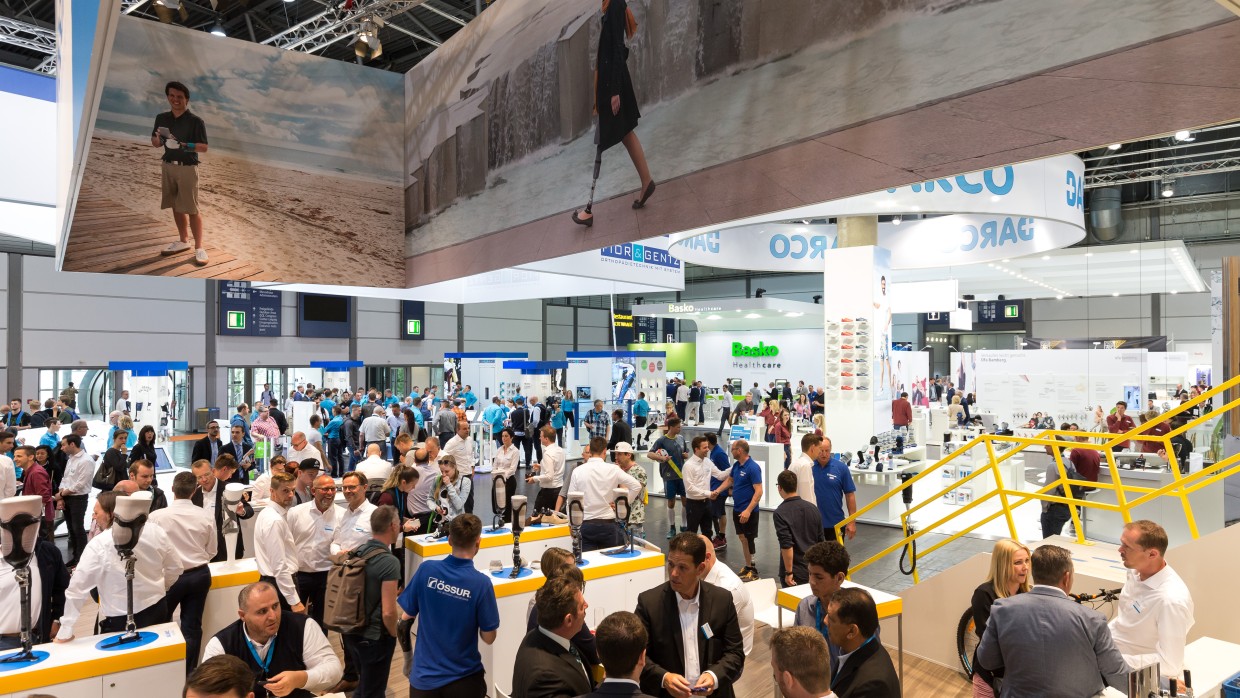 Exhibition booth at OTWorld with numerous visitors, various exhibits such as leg prostheses on tables, and large ceiling banners showing people with prostheses; in the background, booths of companies like “DARCO,” “Basko,” and “Össur” are visible. Exhibition booth at OTWorld with numerous visitors, various exhibits such as leg prostheses on tables, and large ceiling banners showing people with prostheses; in the background, booths of companies like “DARCO,” “Basko,” and “Össur” are visible.