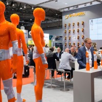 SPORLASTIC booth at OTWorld featuring numerous orange full-body dummies wearing orthoses on arms, legs, and torso; in the foreground, two people are talking at a standing table, while others are seated at tables in the background, and a large screen displays a product image. SPORLASTIC booth at OTWorld featuring numerous orange full-body dummies wearing orthoses on arms, legs, and torso; in the foreground, two people are talking at a standing table, while others are seated at tables in the background, and a large screen displays a product image.