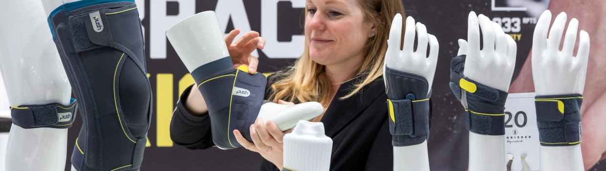 A woman demonstrates an ankle brace on a white model leg with a yellow sports shoe at the OTWorld booth; on the table are additional model arms and legs fitted with gray braces by the brand PUSH, with the text “ANKLE BRACE” visible in the background. A woman demonstrates an ankle brace on a white model leg with a yellow sports shoe at the OTWorld booth; on the table are additional model arms and legs fitted with gray braces by the brand PUSH, with the text “ANKLE BRACE” visible in the background.
