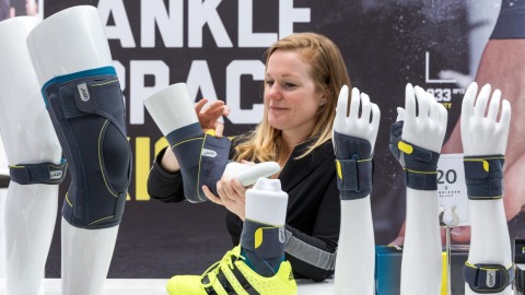 A woman demonstrates an ankle brace on a white model leg with a yellow sports shoe at the OTWorld booth; on the table are additional model arms and legs fitted with gray braces by the brand PUSH, with the text “ANKLE BRACE” visible in the background.