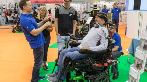 A man in a powered wheelchair with a special head control system is talking to several people at OTWorld, including two men in blue polo shirts and a younger man; in the background, more wheelchairs, informational material, and a screen are visible.