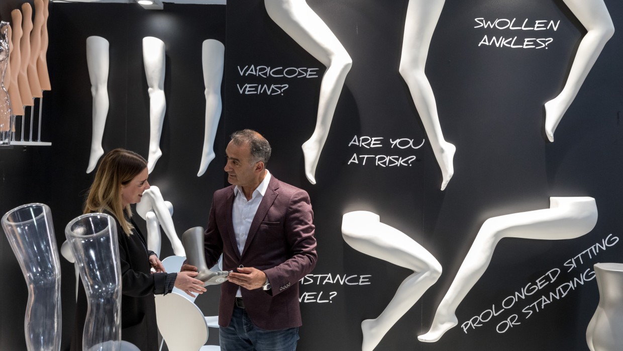 Two people stand at OTWorld in front of a black display wall with white model legs and large text such as “VARICOSE VEINS?” and “PROLONGED SITTING OR STANDING?”; they are examining a leg-shaped exhibit together. Two people stand at OTWorld in front of a black display wall with white model legs and large text such as “VARICOSE VEINS?” and “PROLONGED SITTING OR STANDING?”; they are examining a leg-shaped exhibit together.