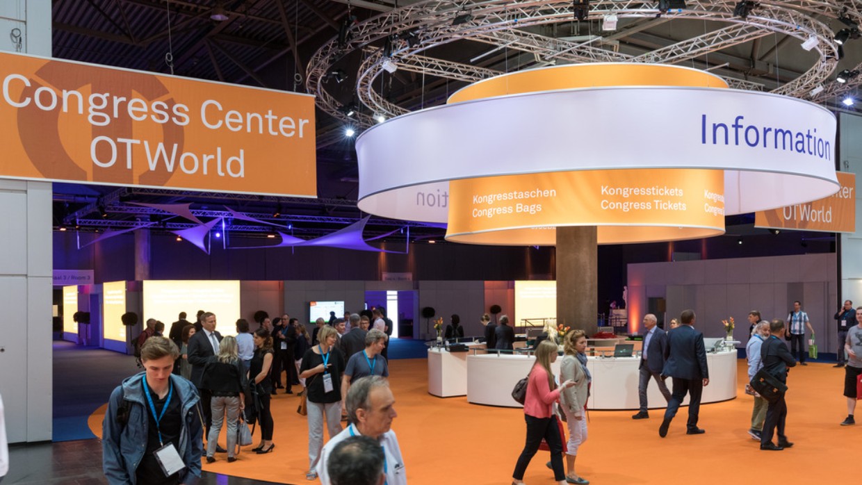 Visitors walk through the Congress Center at OTWorld, featuring an orange floor and a large circular information booth above which hangs signage for congress bags and tickets; to the left, a large banner reads “Congress Center OTWorld.” Visitors walk through the Congress Center at OTWorld, featuring an orange floor and a large circular information booth above which hangs signage for congress bags and tickets; to the left, a large banner reads “Congress Center OTWorld.”