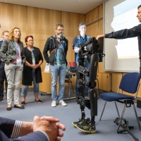 A man demonstrates an exoskeleton at OTWorld in a seminar room to a group of standing attendees; the device is black and fitted with yellow athletic shoes, and a presentation slide is visible on a projection screen in the background. A man demonstrates an exoskeleton at OTWorld in a seminar room to a group of standing attendees; the device is black and fitted with yellow athletic shoes, and a presentation slide is visible on a projection screen in the background.