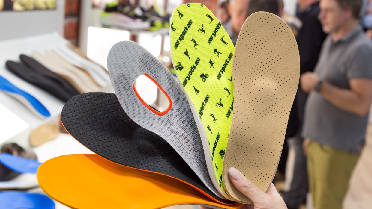 A hand holds five differently designed insoles fanned out at OTWorld; in the background, more insoles are laid out on tables and people are seen in conversation. A hand holds five differently designed insoles fanned out at OTWorld; in the background, more insoles are laid out on tables and people are seen in conversation.