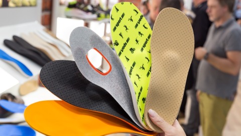 A hand holds five differently designed insoles fanned out at OTWorld; in the background, more insoles are laid out on tables and people are seen in conversation.
