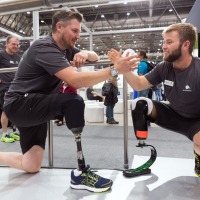 Two men with leg prostheses in sportswear kneel facing each other at OTWorld and greet with a high five; both wear different prosthetic models to demonstrate them to the audience. Two men with leg prostheses in sportswear kneel facing each other at OTWorld and greet with a high five; both wear different prosthetic models to demonstrate them to the audience.