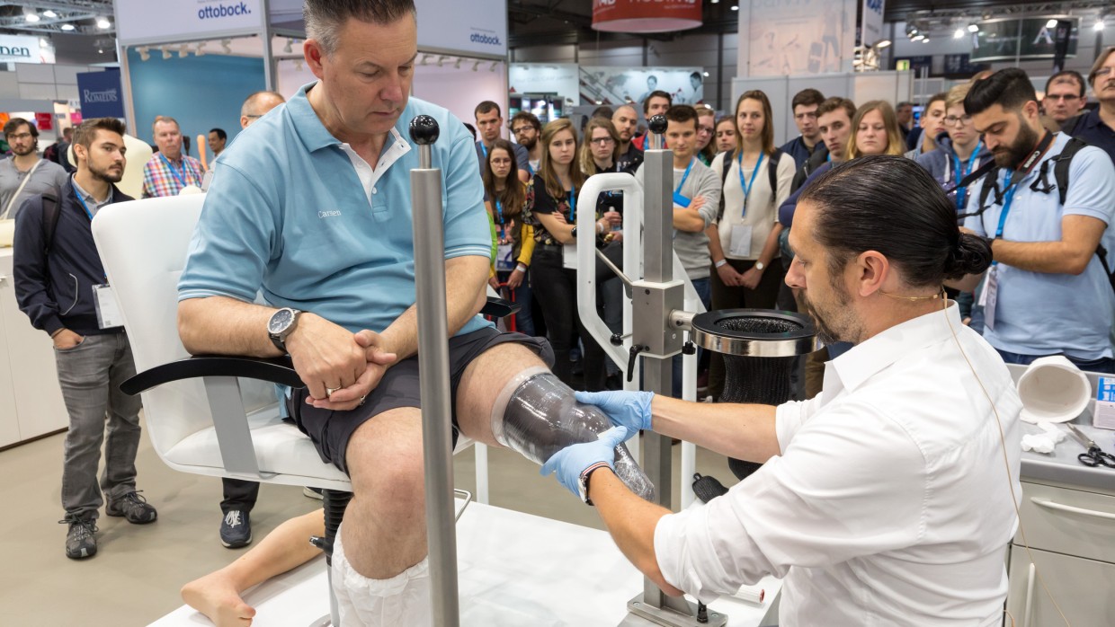 A man sits on a chair at OTWorld while an orthopedic technician fits a transparent mold for a leg prosthesis; numerous trade fair visitors are observing the demonstration in the background. A man sits on a chair at OTWorld while an orthopedic technician fits a transparent mold for a leg prosthesis; numerous trade fair visitors are observing the demonstration in the background.