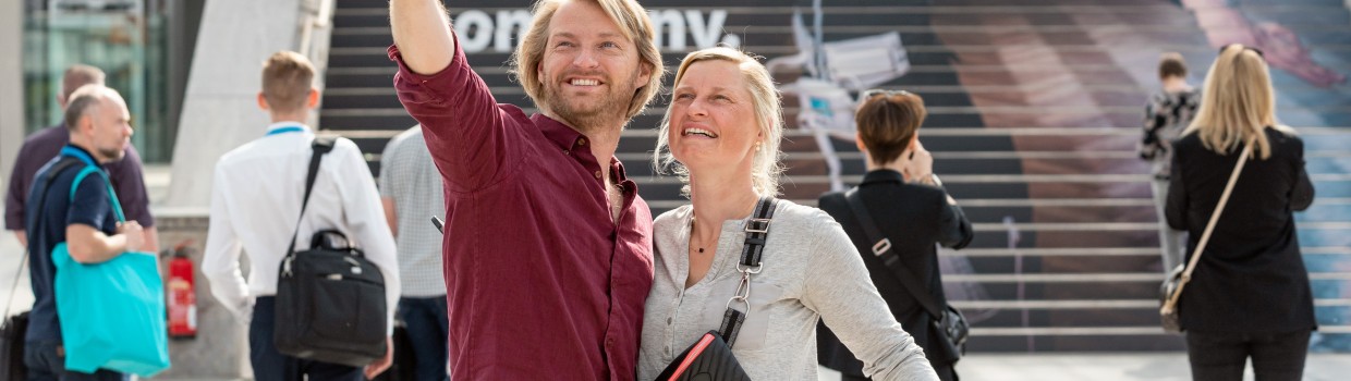 A couple takes a selfie at the OTWorld trade fair; in the background, a large staircase advertisement displays the slogan “The human empowerment company.”