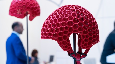 Red protective helmet with honeycomb structure in the foreground, displayed at OTWorld, with trade fair visitors in the background.