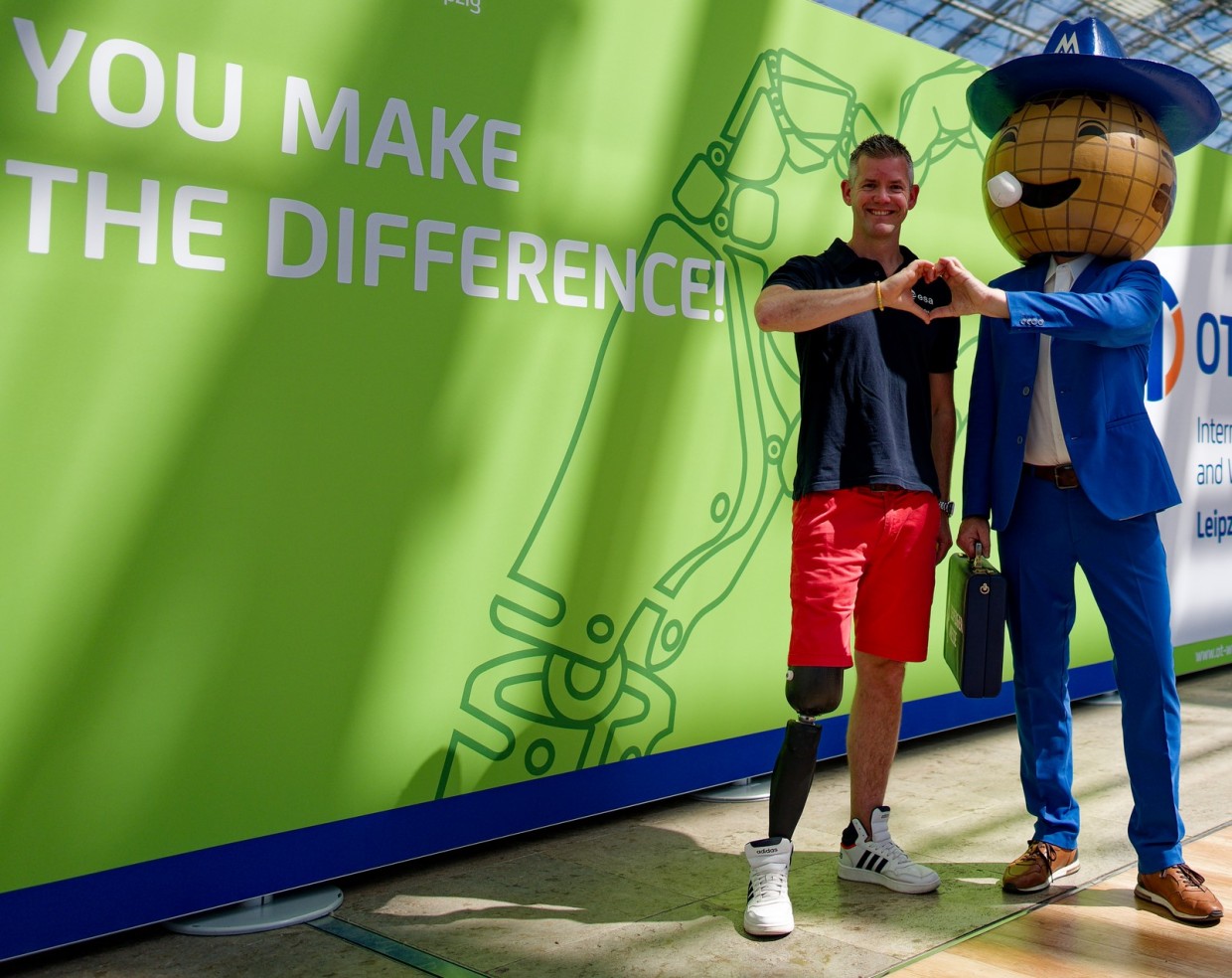 A man wearing red shorts, a black T-shirt, and a prosthetic leg stands next to the Messemännchen, the mascot of the Leipzig Trade Fair, dressed in a blue suit, brown shoes, and a large globe-shaped head with a hat; both form a heart shape with their hands in front of a green wall with the text “YOU MAKE THE DIFFERENCE!”.