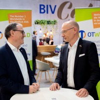 Two men are talking at a bar table. The OTWorld logo can be seen on adisplay in the background. Two men are talking at a bar table. The OTWorld logo can be seen on adisplay in the background.