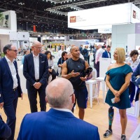 Group of people gathered at the OT-World trade fair around two athletic individuals with prostheses, engaged in lively discussion within a modern exhibition area. Group of people gathered at the OT-World trade fair around two athletic individuals with prostheses, engaged in lively discussion within a modern exhibition area.
