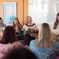 Four women on a panel speaking during a presentation, one woman standing and actively moderating in front of an audience. Four women on a panel speaking during a presentation, one woman standing and actively moderating in front of an audience.
