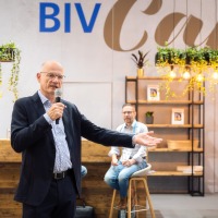 Man speaking into a microphone at the BIV Café during OTWorld, with a cozy café setting in the background including bar stools, plants, and shelves Man speaking into a microphone at the BIV Café during OTWorld, with a cozy café setting in the background including bar stools, plants, and shelves