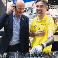 Man with prosthetic arm explains DJ equipment while another man with headphones listens and smiles. Man with prosthetic arm explains DJ equipment while another man with headphones listens and smiles.