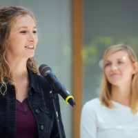 A woman speaks into the microphone. Another female person stands in the background. A woman speaks into the microphone. Another female person stands in the background.