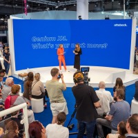 A stage with two speakers in front of a blue back wall, with a large audience and a cameraman in front of it. A stage with two speakers in front of a blue back wall, with a large audience and a cameraman in front of it.