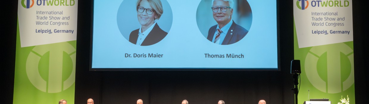 Panel discussion at OT-World with seven experts on stage, their conversation displayed on a large screen, surrounded by OT-World banners.