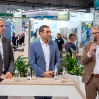 Three men in business attire, one speaking into a microphone, with exhibition booths and trade fair activity in the background. Three men in business attire, one speaking into a microphone, with exhibition booths and trade fair activity in the background.