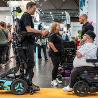 Two men in advanced electric wheelchairs conversing at OT-World, surrounded by other visitors and assistive device booths. Two men in advanced electric wheelchairs conversing at OT-World, surrounded by other visitors and assistive device booths.