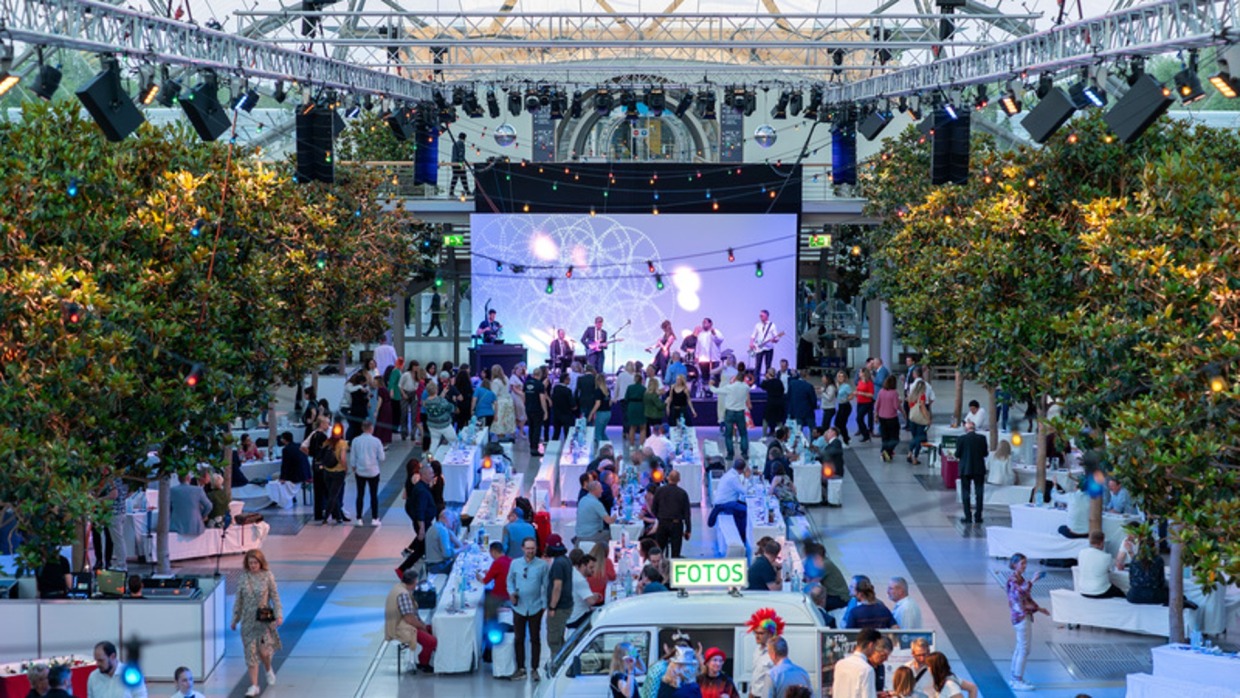 The glass hall of the Leipzig Trade Fair, decorated with trees and lights, is filled with people; in the background, a live band performs on a stage with an LED screen, in front are tables, a small white vehicle with the sign “FOTOS,” and many guests in festive attire.