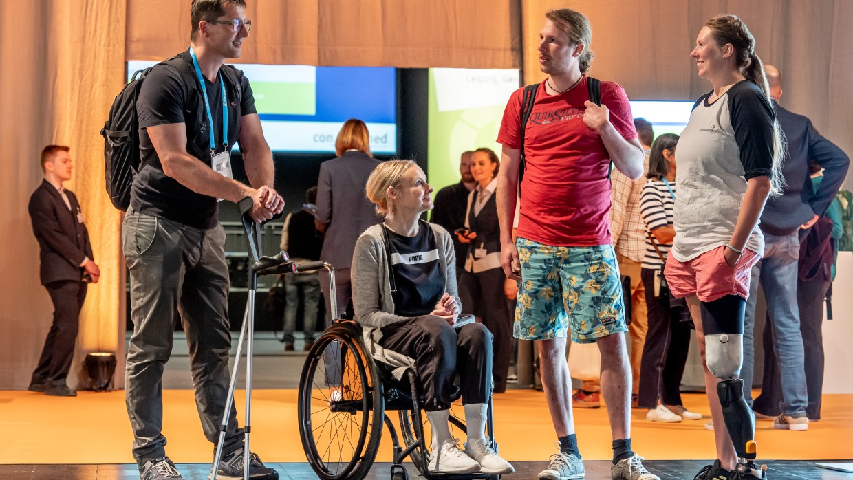 Four visitors with mobility aids and prostheses engage in conversation in a relaxed atmosphere at OTWorld. Four visitors with mobility aids and prostheses engage in conversation in a relaxed atmosphere at OTWorld.